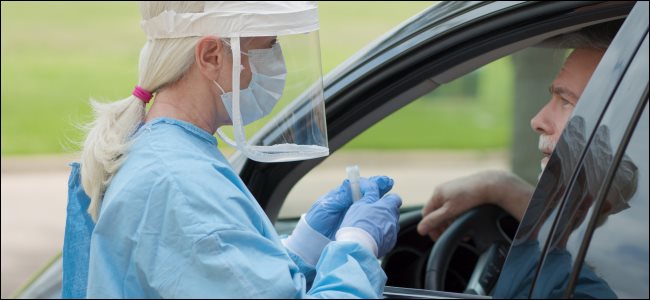 Una mujer con una máscara completa, bata médica y guantes sosteniendo un frasco y hablando con un hombre sentado en un automóvil.