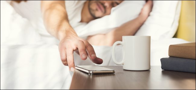 Un hombre soñoliento en la cama buscando un teléfono inteligente en una mesita de noche.