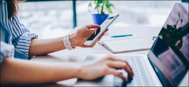 La mano de una mujer que sostiene un teléfono inteligente mientras escribe en un teclado MacBook con el otro.