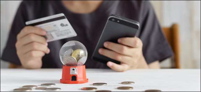 Las manos de un niño sosteniendo una tarjeta de crédito y un teléfono inteligente, y un juego de gacha en la mesa.