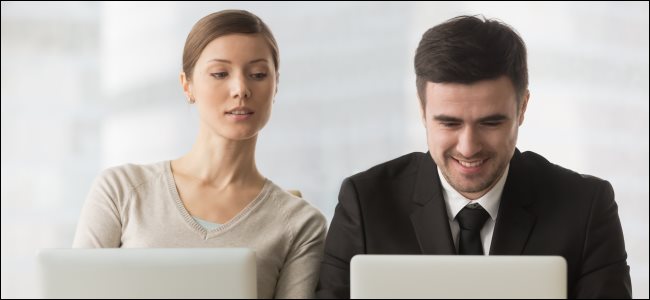 Una mujer mirando por encima del hombro de un compañero de trabajo y espiando mientras usa una computadora portátil.