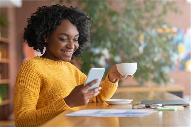 Mujer tomando café y sonriendo mientras mira su teléfono inteligente
