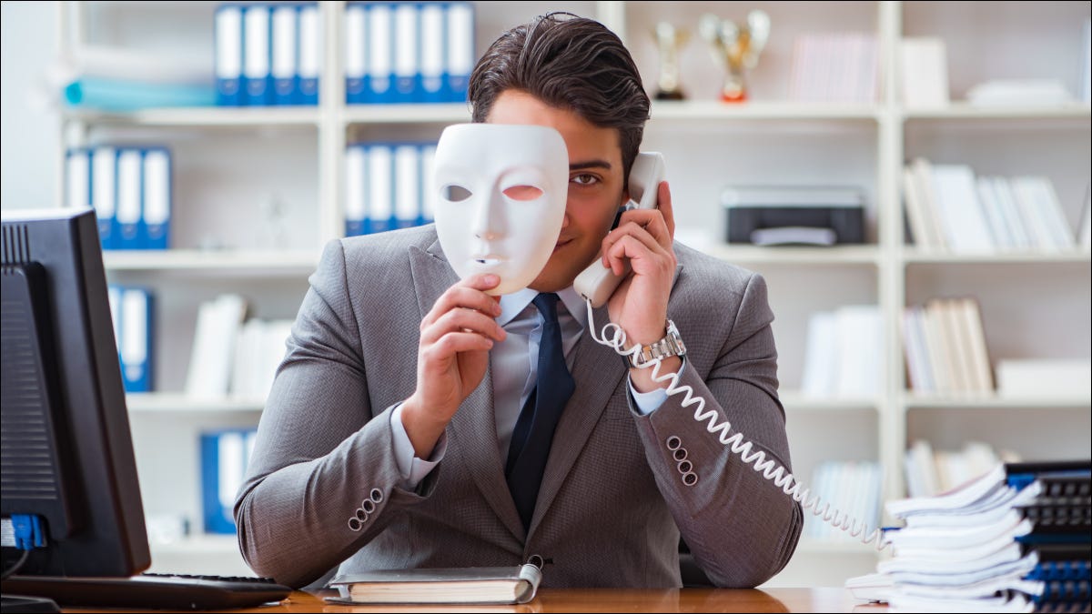 Un hombre de negocios en una oficina haciendo una llamada telefónica y asomándose por detrás de una máscara facial.