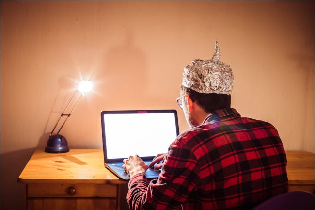 Joven sentado en un escritorio con una computadora portátil y un sombrero de papel de aluminio.