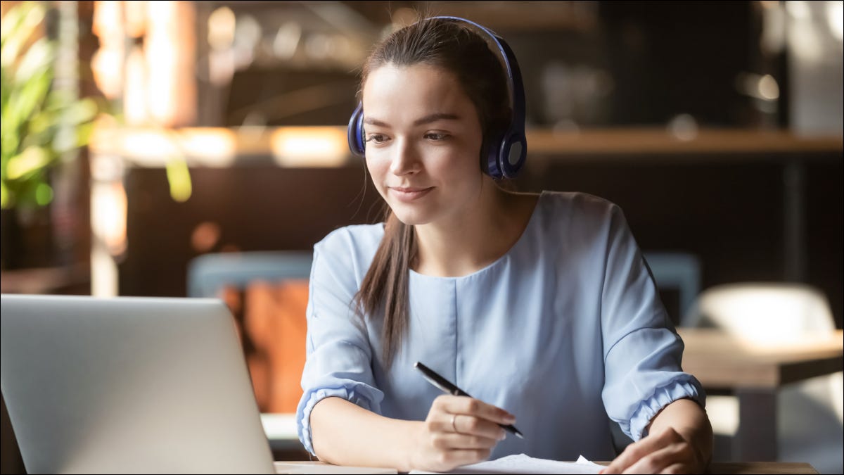 Mujer joven con auriculares mientras toma notas frente a una computadora portátil,
