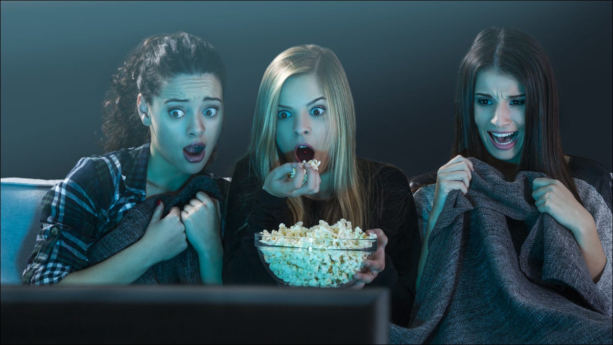Tres mujeres jóvenes viendo una película y comiendo palomitas de maíz con expresiones asustadas.