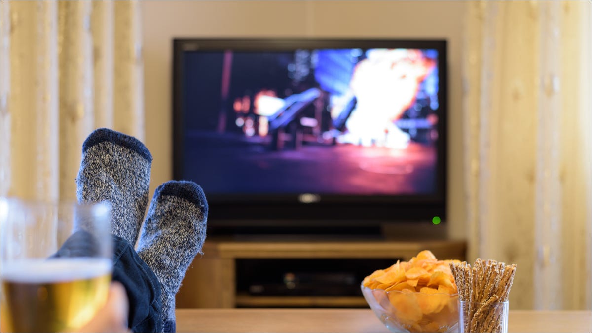 Los pies y bocadillos de una persona en una mesa de café frente a un televisor con una película de acción en exhibición.