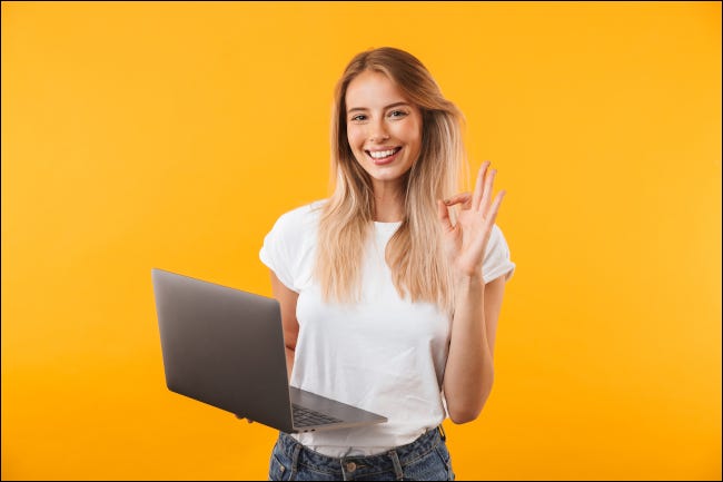 Una mujer joven sonriendo y dando un gesto de "bien" con la mano mientras sostiene una computadora portátil.