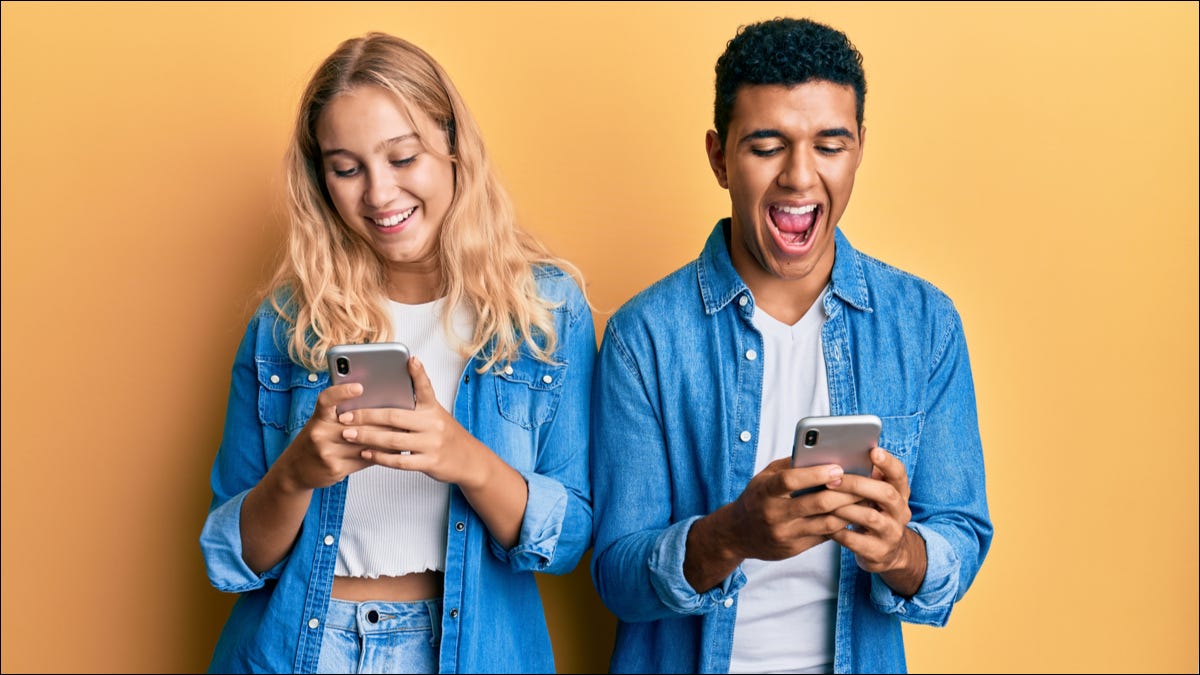 Una pareja joven parada una al lado de la otra mientras sonríe y usa sus teléfonos inteligentes.